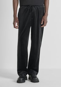 Zwarte fluwelen broek met een losse pasvorm, elastische tailleband en trekkoord. De stof heeft een gladde textuur en combineert goed met stevige zwarte schoenen.