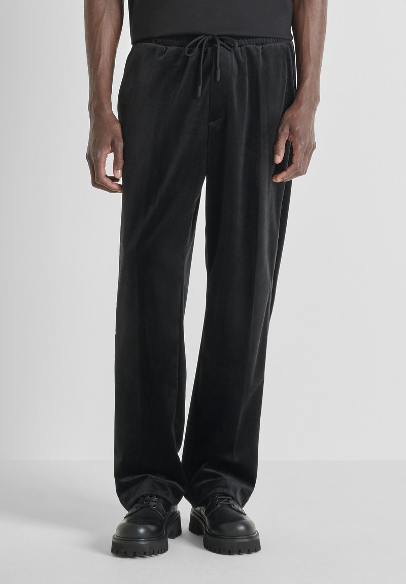 Zwarte fluwelen broek met een losse pasvorm, elastische tailleband en trekkoord. De stof heeft een gladde textuur en combineert goed met stevige zwarte schoenen.