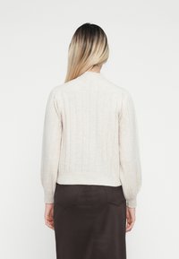 Vaaleanbeige ribattu neulepaita, jossa on pitkät, hieman puffatut hihat ja pyöreä kaulus, puettuna tummanruskean hameen päällä.