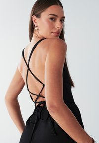 Zwarte jumpsuit met een kruiselings rugontwerp met verstelbare strikjes, smalle bandjes en een gladde stoftextuur.