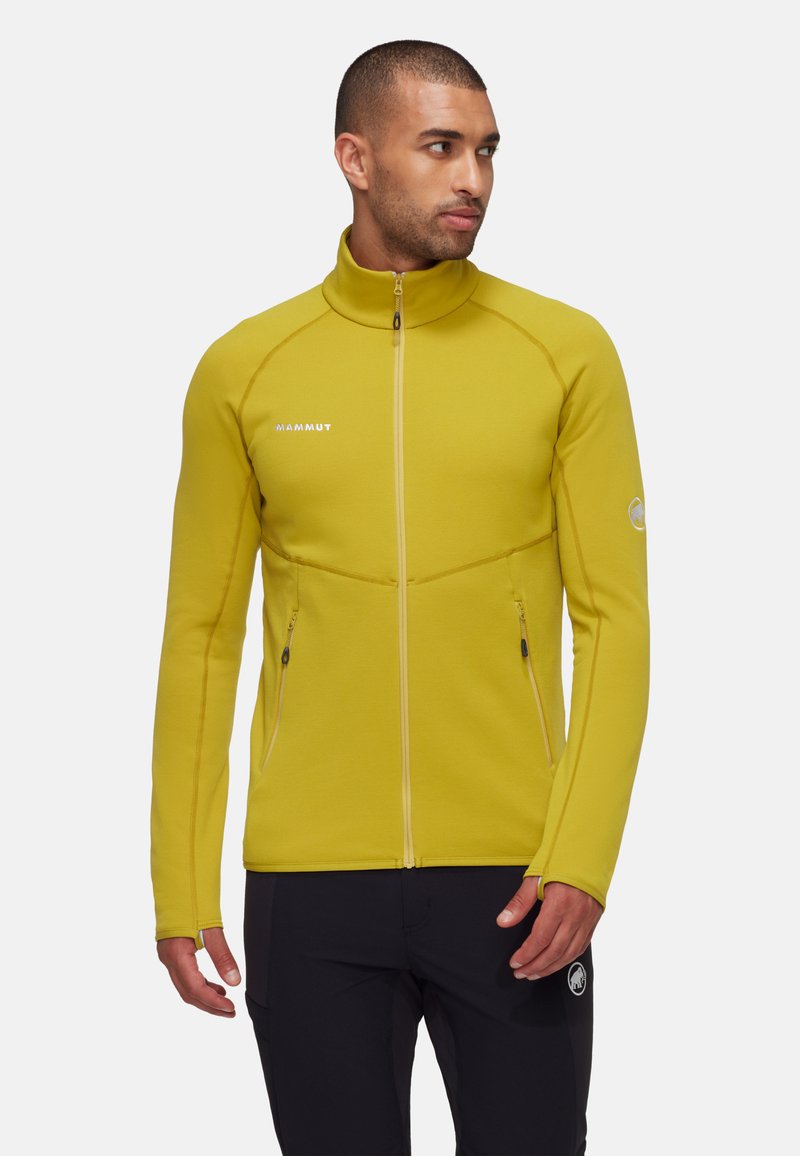 Geel fleece jack met ritssluiting en hoge kraag, strakke pasvorm, zijzakken en contrasterende ritsdetails, gecombineerd met donkere broek.