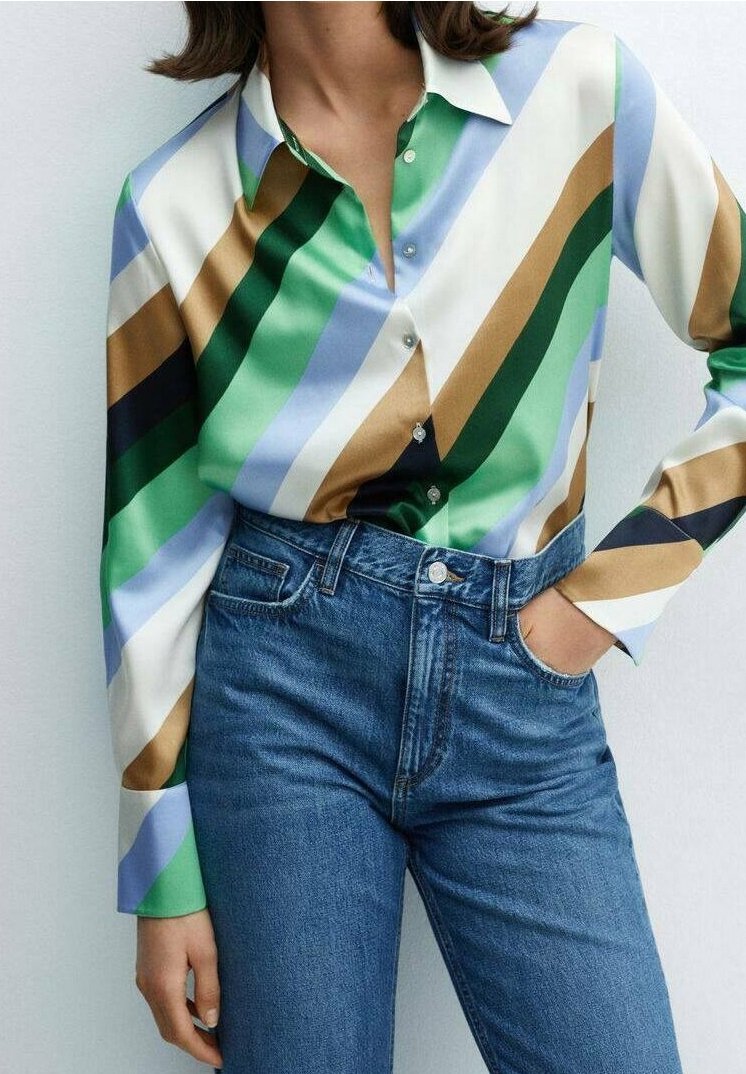 Frau in einer langärmligen Seidenbluse mit diagonalen Streifen in Grün, Blau, Schwarz, Weiß und Beige, kombiniert mit hochgeschnittenen blauen Jeans.