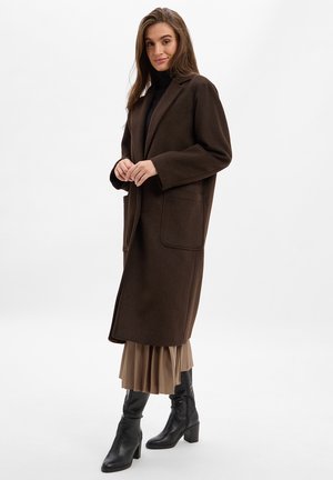 Bruine wollen jas met een ontspannen pasvorm, grote Franse zakken en notch-revers, gecombineerd met een beige geplooide rok en zwarte haklaarzen.