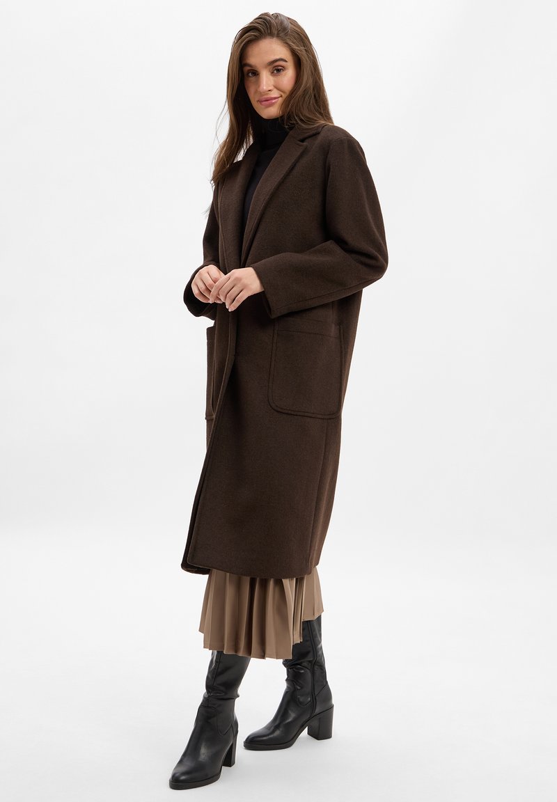 Brauner Wollmantel in lockerer Passform, mit großen Fronttaschen und Reverskragen, kombiniert mit einem beigen plissierten Rock und schwarzen Stiefeln mit Absatz.