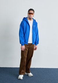 Blauwe rits-hoodie met voorzakken, gecombineerd met een beige t-shirt en bruine broek. Accessoires zijn onder andere oranje getinte zonnebril en witte sneakers.