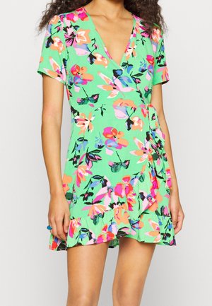 Femme portant une robe portefeuille verte à manches courtes avec un motif floral coloré et un ourlet à volants, posant devant un fond uni.