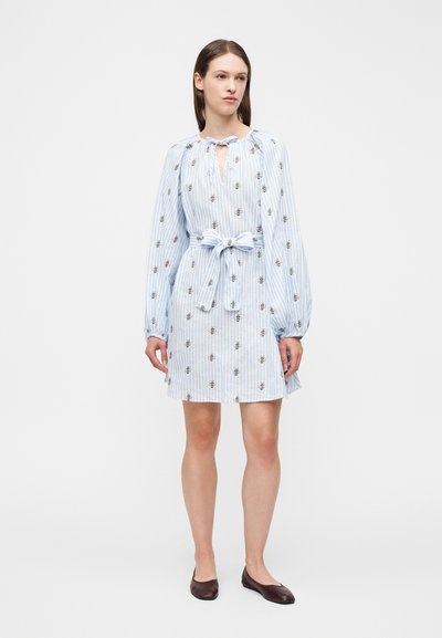 Γυναίκα που φοράει ανοιχτό μπλε φόρεμα με ρίγες και σχέδια από μέλισσες, με φιόγκο στη μέση, μακριά μανίκια και σκοτεινά επίπεδα παπούτσια, στέκεται μπροστά σε απλό λευκό φόντο.