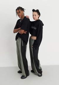 Two individuals stand close together, one in a black oversized t-shirt and black pants with green accents, the other in a black cropped hoodie and similar pants.