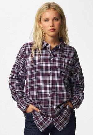 Une femme blonde pose, les mains dans les poches de son jean. Elle porte une chemise boutonnée à carreaux bordeaux et blancs, sur un fond blanc uni.