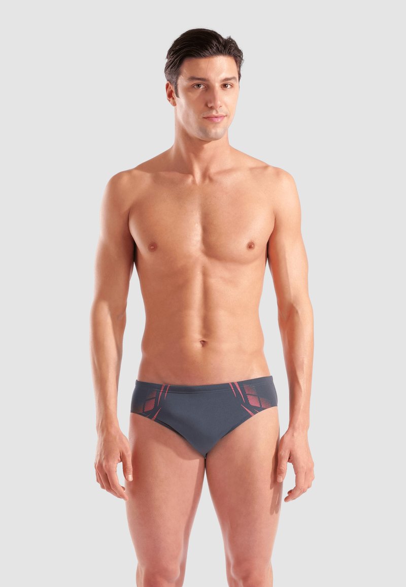 Giovane uomo muscoloso in piedi di fronte, indossando slip da bagno scuri con motivi geometrici rossi su uno sfondo chiaro e semplice.