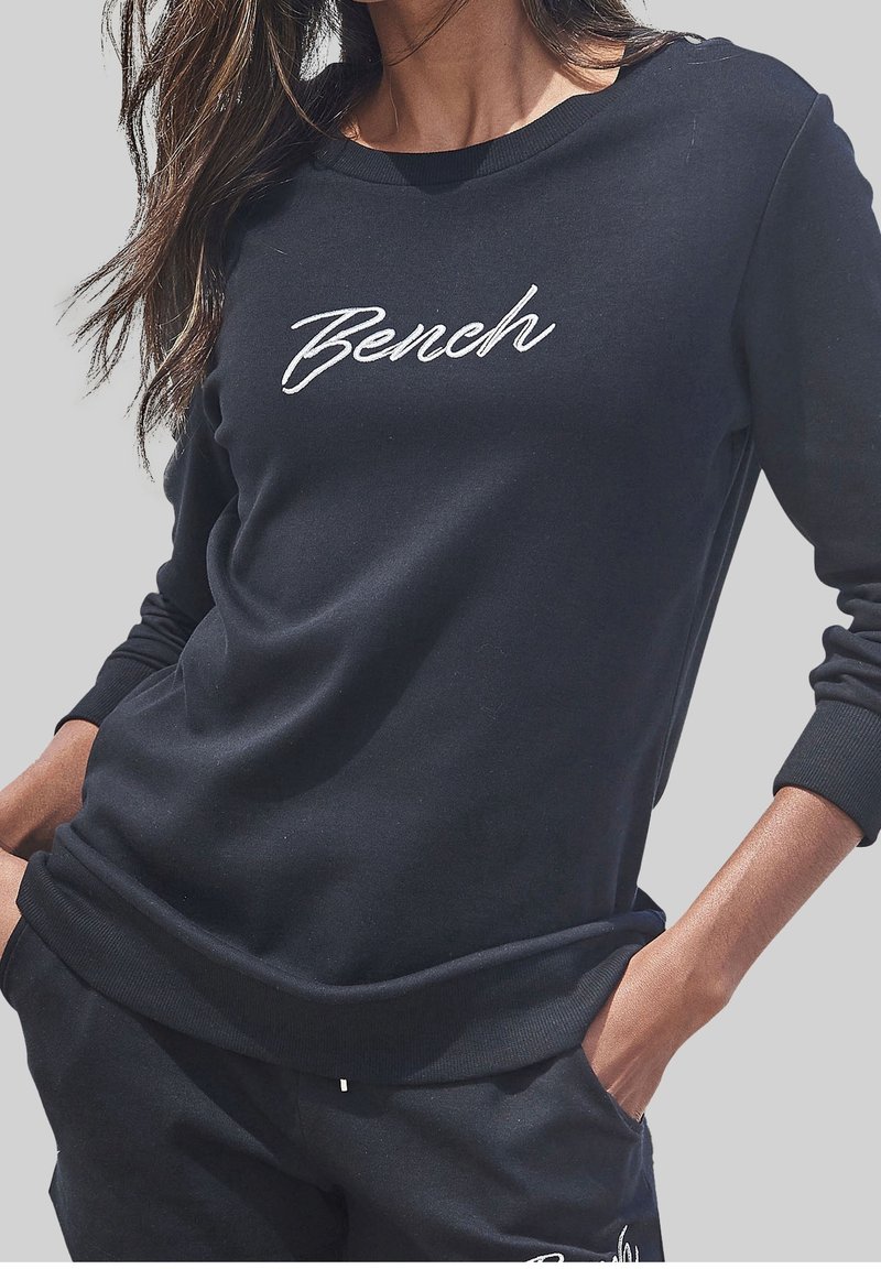 Femme portant un sweat-shirt noir à manches longues avec « Bench » écrit en blanc sur la poitrine, les mains dans les poches du pantalon noir assorti.