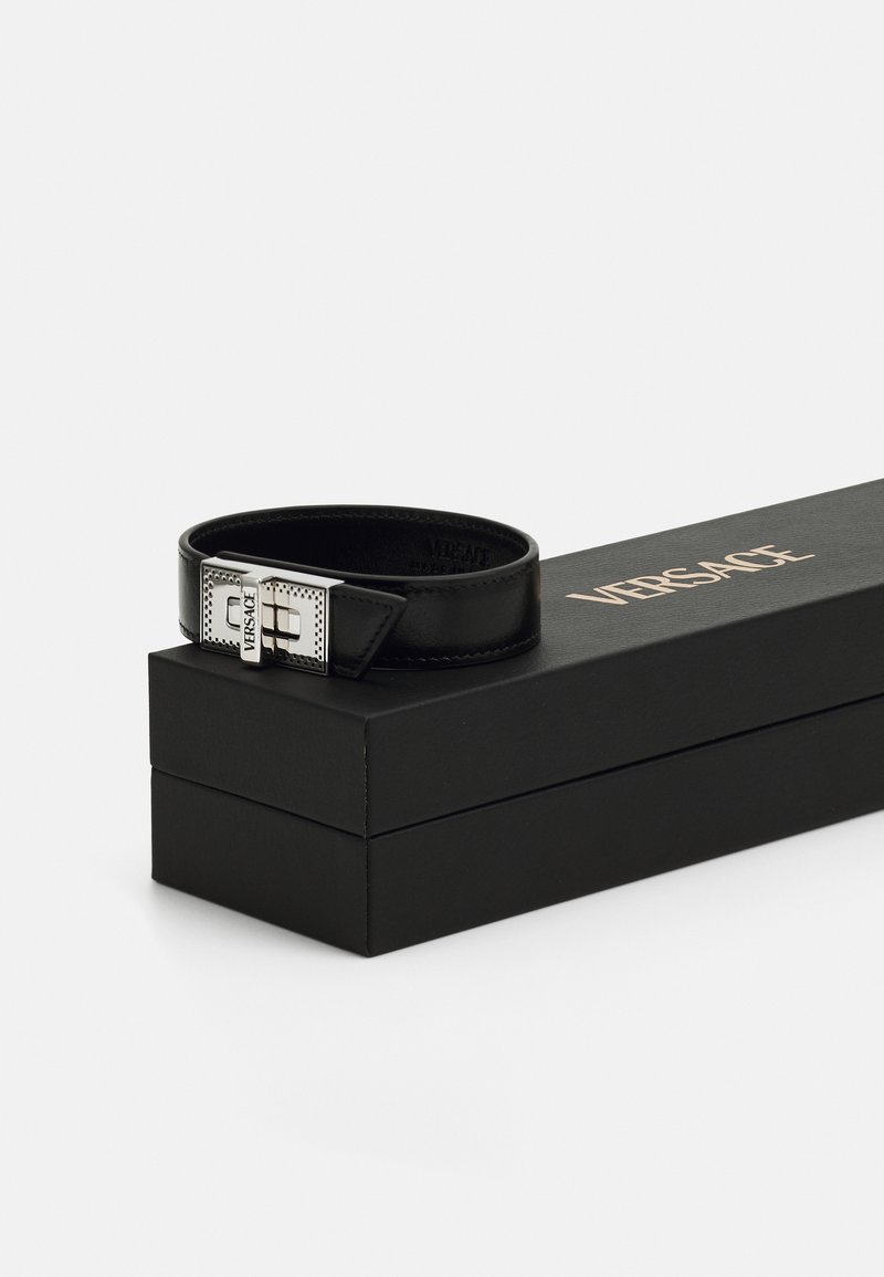 Schwarzer Leder-Gürtel mit einer silbernen Schnalle, die ein durchbrochenes Design aufweist, präsentiert in einer schwarzen Box mit goldener "VERSACE"-Beschriftung.