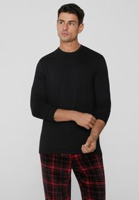 Černé tričko s dlouhým rukávem a kulatým výstřihem, doplněné červenými a černými kostkovanými pyžamovými kalhotami. Tkanina vypadá hladce a má přiléhavý střih.