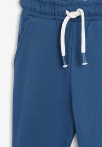 Blå bomulds sweats med elastisk talje, hvid snor med blå spidser og en synlig sidelomme på venstre ben.