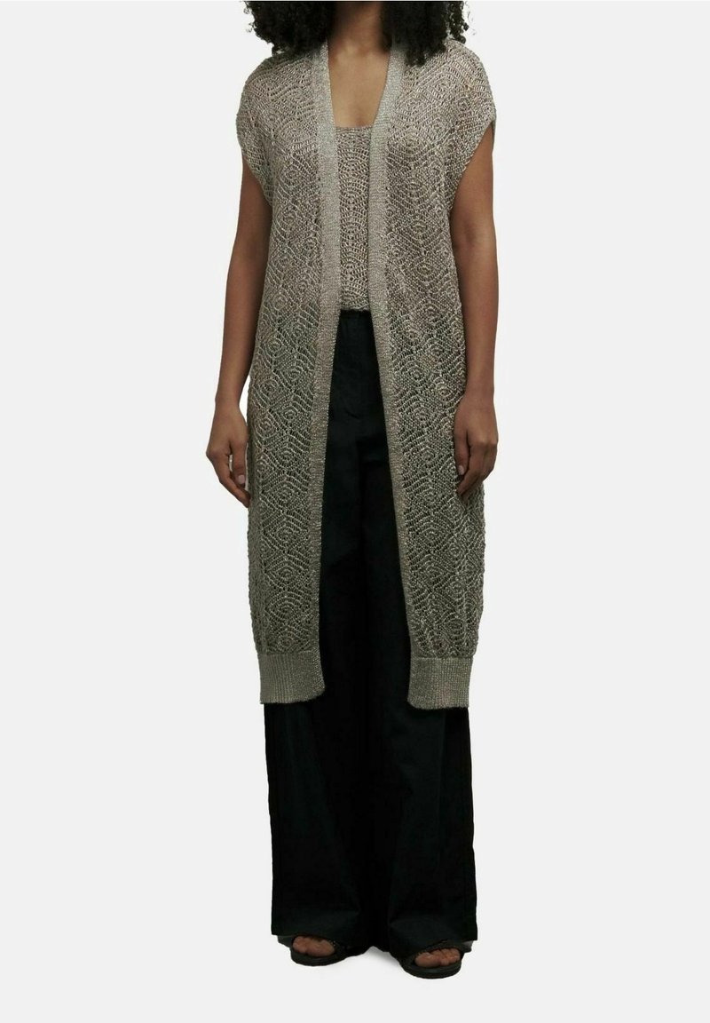 Femme portant un cardigan long en tricot beige sans manches, par-dessus un haut assorti et un pantalon noir large, ainsi que des sandales à bouts ouverts.