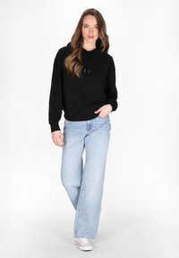 Felpa nera con cappuccio e lacci, maniche ampie e vestibilità rilassata, abbinata a jeans larghi blu chiaro e sneakers bianche.