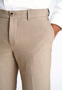 Pantaloni beige su misura con una texture liscia, gamba dritta e chiusura con un bottone in vita. Presentano tasche laterali e passanti per la cintura.