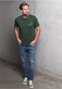 Groene t-shirt met korte mouwen en een zakdetail, gecombineerd met lichtblauwe jeans en groene sneakers, staande tegen een betonnen muur.