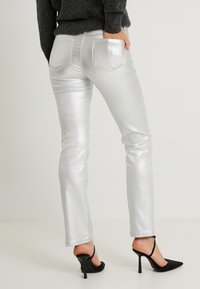 Pantalones vaqueros metálicos plateados de corte ajustado, diseño de cinco bolsillos y costuras en tono dorado, combinados con zapatos negros de tacón alto.