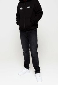 Uomo che indossa una felpa nera con zip e loghi bianchi, jeans scuri e sneakers bianche, in piedi su uno sfondo bianco semplice.