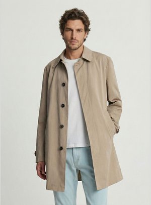 Mężczyzna w beżowym płaszczu zapinanym na guziki, na białej koszulce i jasnoniebieskich dżinsach, stojący na jednolitym jasnym tle.