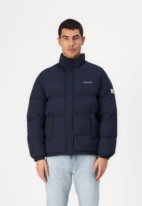 Marineblauwe gewatteerde jas met hoge kraag, voorrits en twee zijzakken. Heeft een logo op de borst en elastische boorden. Vormgevende pasvorm.