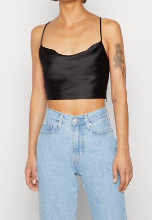 Mujer con una camiseta corta de satén negro con tirantes finos, vaqueros de tiro alto azul claro y dos tatuajes florales en sus brazos superiores.