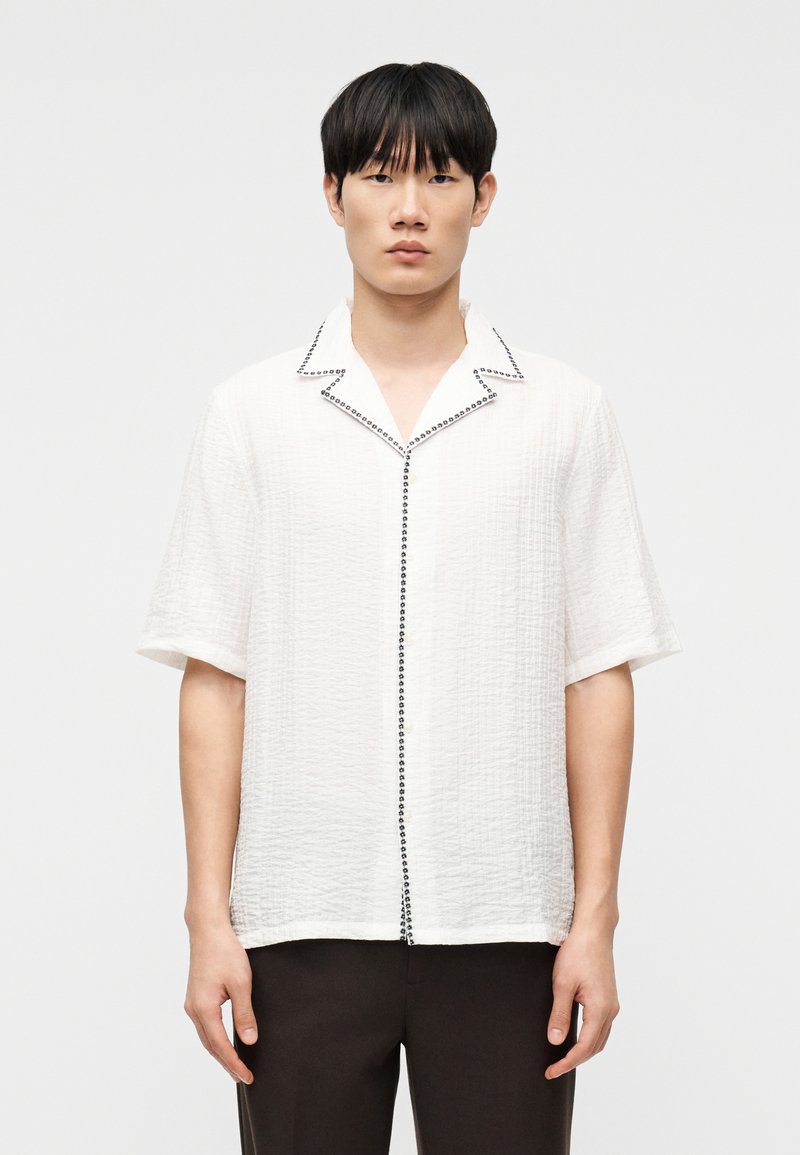 Νεαρός άνδρας με ίσια μαύρα μαλλιά που φοράει λευκό κοντομάνικο υφασμένο πουκάμισο με μαύρο περίγραμμα κατά μήκος του γιακά και των κουμπιών, στέκεται μπροστά σε απλό φόντο.