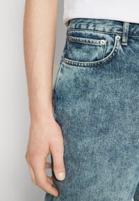 Jeans de mezclilla azul claro con un lavado desgastado, que cuentan con un pequeño bolsillo frontal, acentos de botones metálicos y costuras en contraste. Primer plano del lateral.