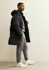 Hombre con abrigo acolchado negro con capucha, pantalones grises de chándal, calcetines blancos y zapatillas blancas, de pie contra una pared lisa y clara.