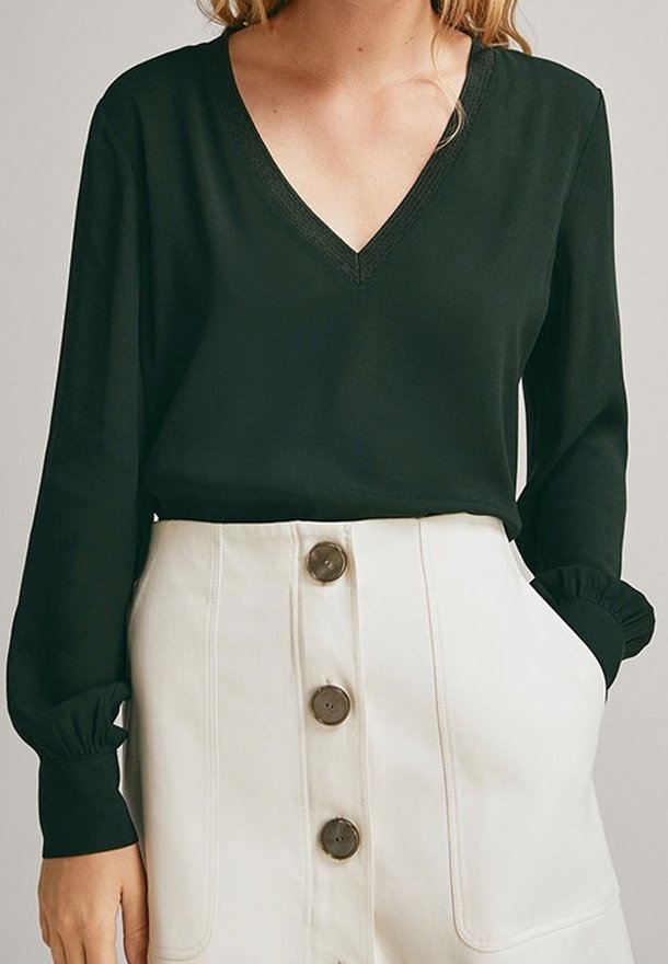 Femme portant un haut noir à manches longues en V, rentré dans une jupe blanche taille haute avec de gros boutons marron à l'avant.