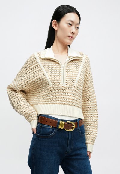 Camisola curta de malha cor creme com fecho frontal de correr e padrão texturizado, combinada com jeans azul escuro e um cinto castanho com fivela dourada.