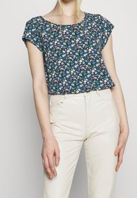 Haut à manches courtes fleuri en bleu marine avec des fleurs blanches, roses et vertes ; coupe ample et encolure arrondie, associé à un pantalon crème.