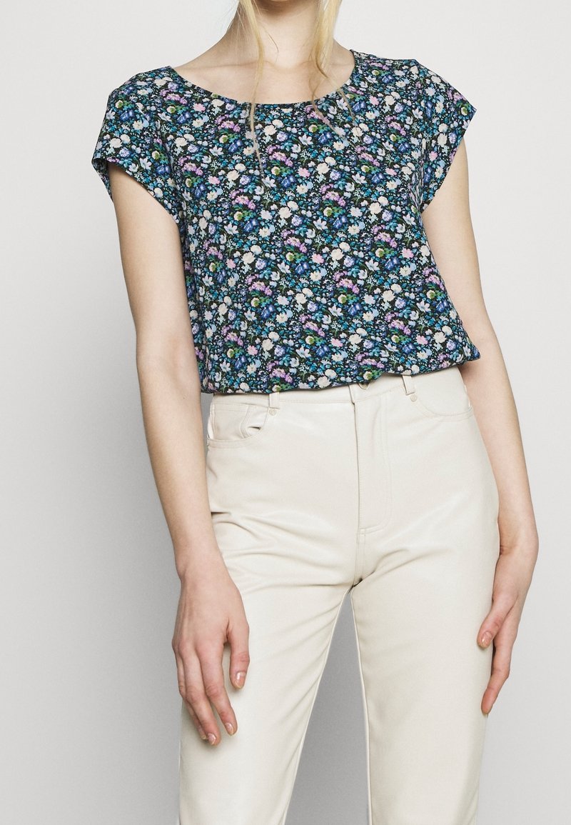 Haut à manches courtes fleuri en bleu marine avec des fleurs blanches, roses et vertes ; coupe ample et encolure arrondie, associé à un pantalon crème.