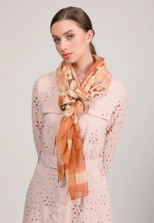 Giovane donna in un vestito rosa pallido con motivo a occhielli che indossa una sciarpa arancione a motivo annodata in modo sciolto intorno al collo, su uno sfondo neutro.