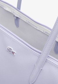 Bolsa tote lila con exterior texturizado de nylon, ribete blanco con marca y un pequeño logo de cocodrilo bordado. Cuenta con asas dobles y cierre con cremallera.