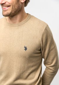 Khaki trui met een geribde halslijn, gemaakt van zachte stof. Voorzien van een klein marineblauw logo op de linkerborst. Soepele textuur met een getailleerd ontwerp.