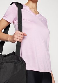 Lichtroze, kortekleurige, v-hals sportshirt gemaakt van een gladde stof. Een zwarte sporttas wordt over de schouder gehouden, met een zichtbare ritsdetail.