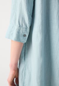 Manche de chemise en lin bleu clair retroussée et boutonnée au-dessus du poignet, avec couture latérale visible et une partie du bras du porteur.
