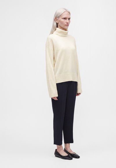 Femme aux cheveux lisses et clairs portant un pull à col roulé crème, un pantalon noir court et des chaussures plates noires, debout de profil sur un fond uni.