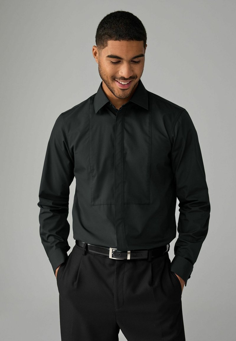 Schwarzes Langarmshirt mit strukturiertem Kragen, featuring ein flaches Frontpanel-Design und Knopfverschluss, kombiniert mit schwarzen texturierten Hosen.