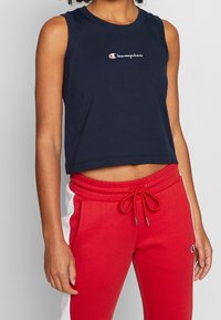 Granatowy top na ramiączkach o krótkim kroju z logo Champion na piersi, w zestawieniu z czerwonymi spodniami dresowymi z białymi panelami po bokach i sznurkiem w pasie.