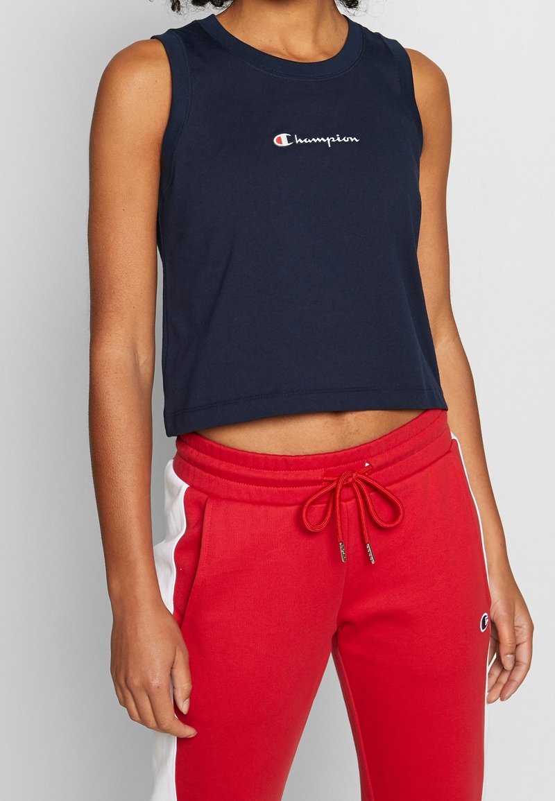 Granatowy top na ramiączkach o krótkim kroju z logo Champion na piersi, w zestawieniu z czerwonymi spodniami dresowymi z białymi panelami po bokach i sznurkiem w pasie.