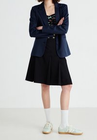 Personne portant un blazer bleu marine, une jupe noire, un haut à fleurs, des chaussettes blanches et des baskets bleu clair, debout les bras croisés sur fond blanc.