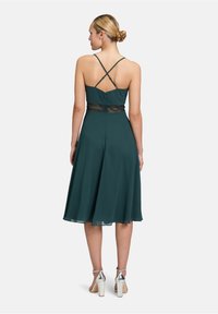 Une robe teal avec une jupe évasée, dotée de bretelles croisées et d'un détail en dentelle à la taille. Le tissu semble léger et doux. Des escarpins argentés sont portés.