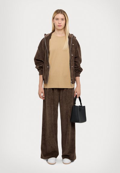 Brown velour zip-up jacket, beige t-shirt, and matching wide-leg pants. Holding a small black handbag. White shoes with light brown soles.