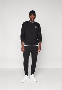 Svart hettegenser med subtil grafikk, kombinert med sorte joggebukser. Hvite detaljer på halsbåndet til hettegenseren. Hvite sneakers fullfører antrekket.