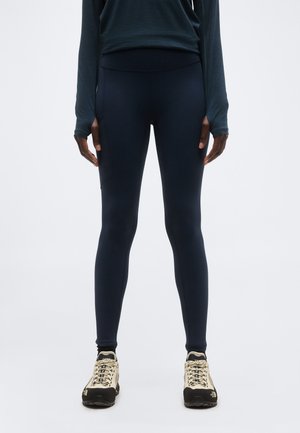 Άτομο που φοράει μαύρο κολάν, σκούρο μακρυμάνικο τοπ και μπεζ πεζοπορικά παπούτσια, στέκεται μπροστά σε απλό φωτεινό γκρι φόντο.