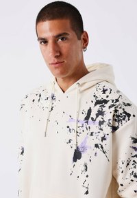 Jonge man met kort haar in een crèmekleurige hoodie bespat met zwarte en paarse verf, die recht in de camera kijkt met een neutrale uitdrukking.