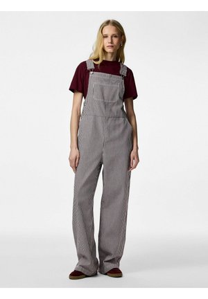 Jeune femme aux longs cheveux blonds portant un t-shirt bordeaux et une salopette ample à rayures, debout devant un fond blanc uni.